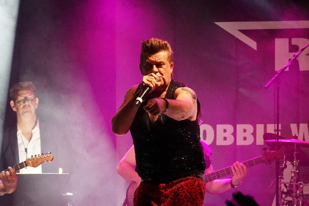 Ein Sänger mit Mikrofon performt auf einer Bühne, umgeben von Nebel und Bandmitgliedern mit Instrumenten.