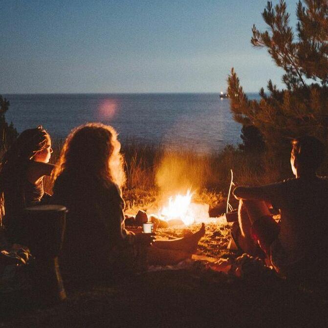 Drei Personen sitzen am Lagerfeuer am Strand, im Hintergrund ist das Meer und der nächtliche Himmel zu sehen.