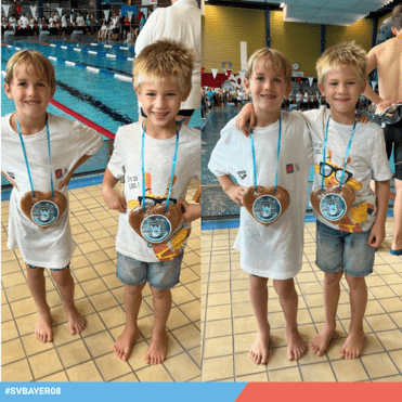 Zwei Jungen in Badeshorts und T-Shirts mit Medaille in Form eines Lebkuchenherzens, Schwimmbecken im Hintergrund.