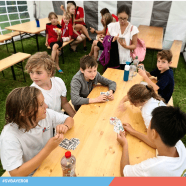 Gruppe von Kindern spielt Karten an einem Tisch im Zelt, einige essen im Hintergrund, während eine Erwachsene beobachtet.