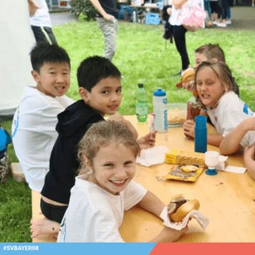 Fünf Kinder sitzen an einem Tisch im Freien, essen Snacks und trinken aus Flaschen. Lächeln und genießen die Zeit.