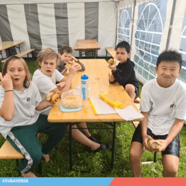 Fünf Kinder sitzen an einem Tisch in einem Zelt, essen Hotdogs und trinken aus einer Wasserflasche.