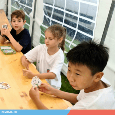 Drei Kinder sitzen an einem Tisch in einem Zelt und spielen Karten mit einem Kartenspiel auf der Holzoberfläche.