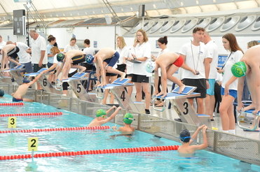 Schwimmer stehen an Startblöcken am Beckenrand, während Trainer und Zuschauer das Wettkampfgeschehen beobachten.