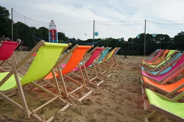 Reihe bunter Liegestühle aus Holz steht im Sand, im Hintergrund ein Volleyballnetz und ein Surfbrett.