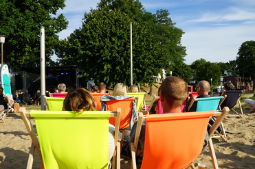 Menschen sitzen in bunten Liegestühlen am Strand und schauen auf eine Bühne mit Lichtinstallationen.