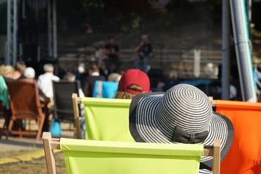 Zuschauer in bunten Liegestühlen, eine Frau mit schwarz-weißem Hut im Fokus, Blick auf eine Bühne im Hintergrund.