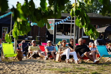 Gruppe von Menschen sitzt entspannt in Liegestühlen an einem Sandstrand, im Hintergrund ein Kiosk unter blauem Himmel.