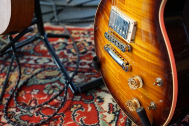 Detailaufnahme einer elektrischen Gitarre auf einem bunten Teppich mit Verstärker und Kabeln im Hintergrund.