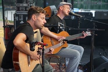 Zwei Musiker mit Akustikgitarren singen in ein Mikrofon auf einer Bühne im Freien, im Hintergrund ein See.