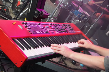 Nahaufnahme eines roten Synthesizers mit Händen, die die Tasten spielen, im Hintergrund ist ein Schlagzeug sichtbar.