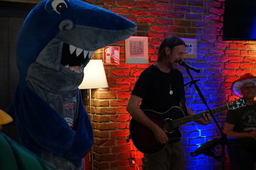 Maskottchen in Hai-Kostüm steht neben einem Sänger mit Gitarre, während im Hintergrund eine rot-blau beleuchtete Wand zu sehen ist.