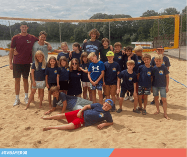 Gruppe von Jugendlichen und Kindern in blauen T-Shirts, posiert am Sandstrand vor einem Beachvolleyballnetz.