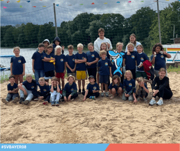 Gruppe von Kindern in blauen T-Shirts steht an einem Strand mit Volleyballnetz und einem See im Hintergrund.