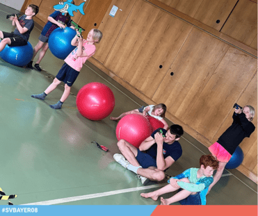 Kinder und ein Erwachsener spielen in einer Halle mit Spielzeugpistolen und Gymnastikbällen.