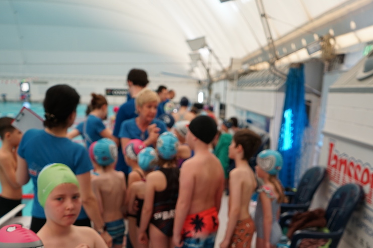 Gruppe von Kindern in Badeanzügen und Badekappen wartet an einem Schwimmbecken, während Trainerin Anweisungen gibt.