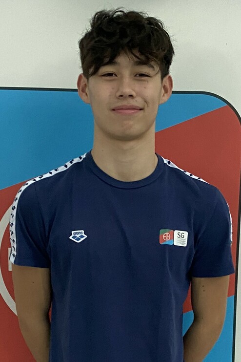 Junger Mann in einem blauen Sportshirt mit Emblem, lächelt vor einer farbigen Wand.