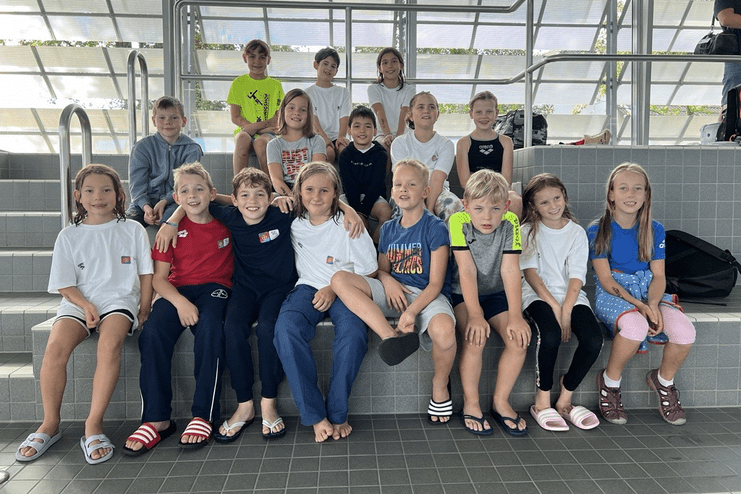 Gruppe von 14 Kindern in Sportbekleidung, sitzend auf Treppen in einem Schwimmbad. Helle, moderne Umgebung.