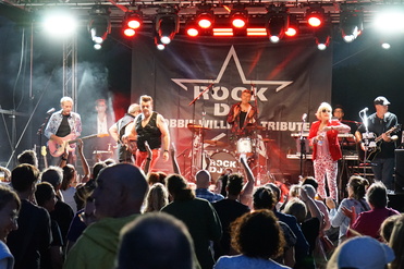 Tribute-Band spielt live auf Bühne, während das Publikum begeistert tanzt und mitfeiert. Beleuchtung in rot und grün.