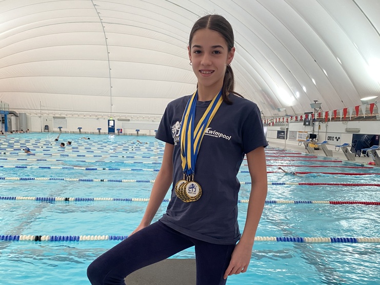 Mädchen mit Medaillen auf einem Startblock in Schwimmhalle, Schwimmbecken im Hintergrund.