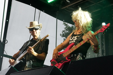 Zwei Musiker auf der Bühne spielen Gitarre und E-Bass, umgeben von Lichtern und einer schwarzen Hintergrundleinwand.