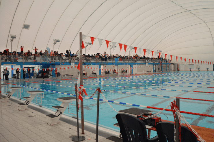 Schwimmhalle mit Wasserbecken, Startblöcken und Zuschauern auf der Tribüne, überdacht von einem Bogen.