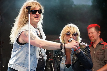 Zwei Sänger auf der Bühne, einer mit Sonnenbrille und langen Haaren, der andere spricht ins Mikrofon.