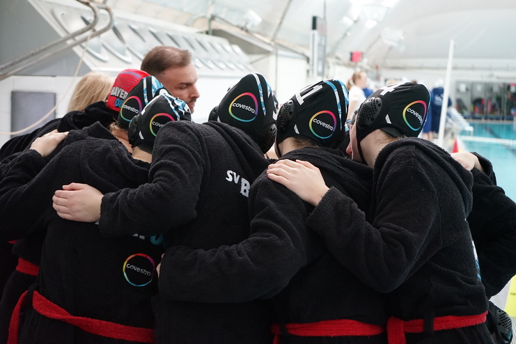 Mädchenteam in schwarz-roten Bademänteln umfängt sich am Beckenrand vor dem Spiel, mit Badekappen auf den Köpfen.