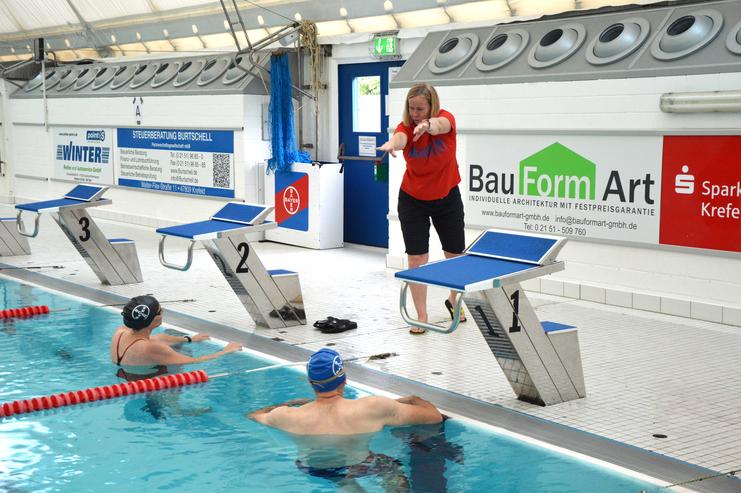 Trainerin gibt Anweisungen an Schwimmer am Beckenrand; Schwimmblöcke und Werbebanner im Hintergrund sichtbar.