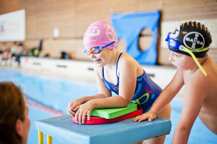 Kind mit Schwimmbrille und Badehaube sitzt auf einem Startblock, vor ihm liegen farbige Schwimmbretter am Beckenrand.