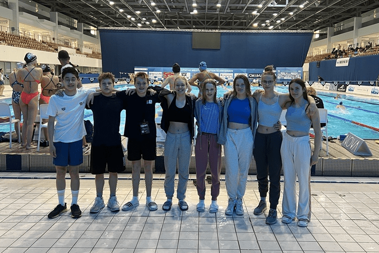 Gruppe von Kindern und Jugendlichen am Beckenrand eines Schwimmwettkampfs in einer Sporthalle.