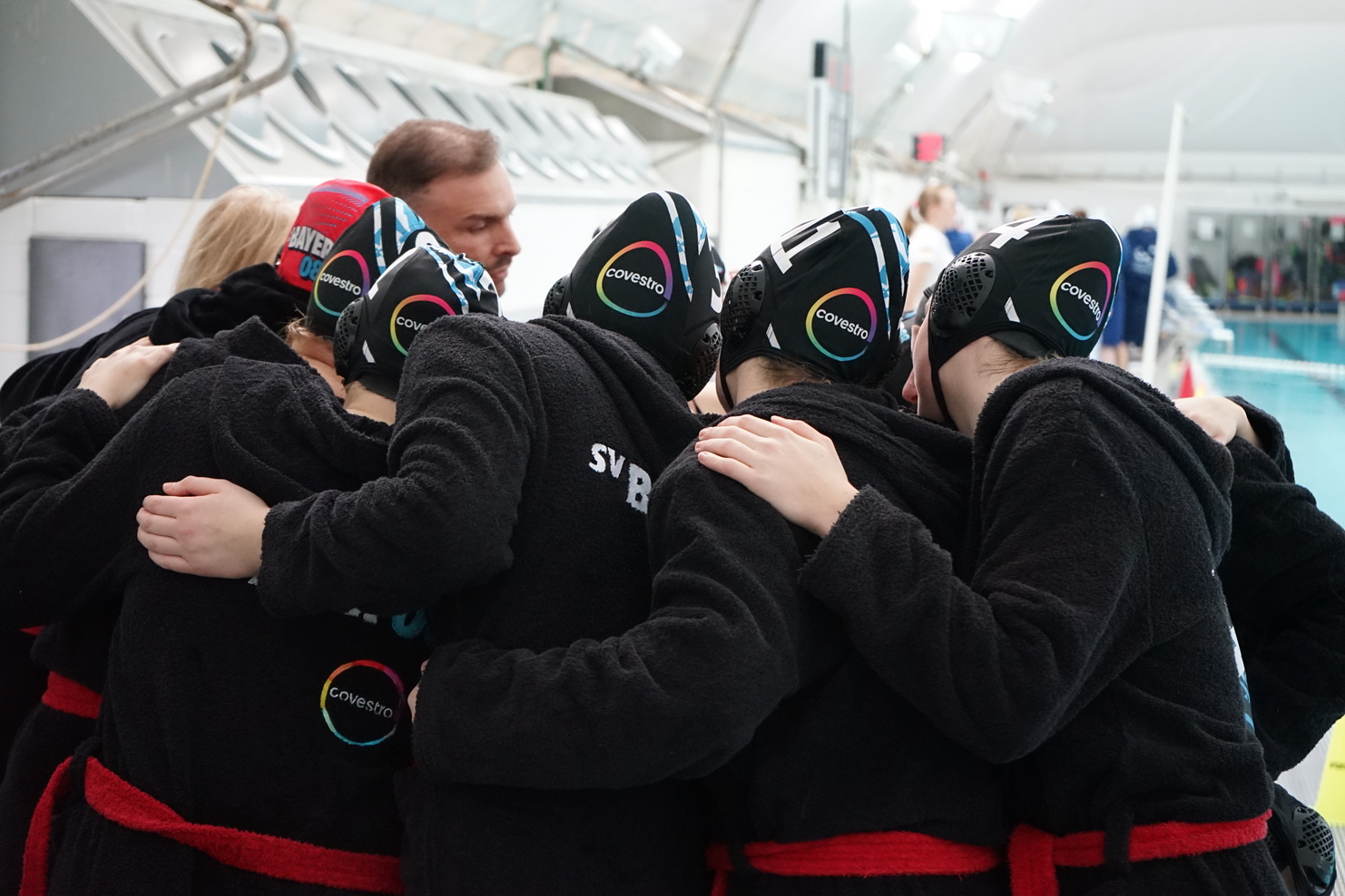 Mädchenteam in schwarz-roten Bademänteln umfängt sich am Beckenrand vor dem Spiel, mit Badekappen auf den Köpfen.
