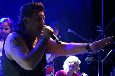 Mann mit tätowiertem Arm singt ins Mikrofon auf einer Bühne, im Hintergrund Kinder und Musiker. Bühnenbeleuchtung ist sichtbar.