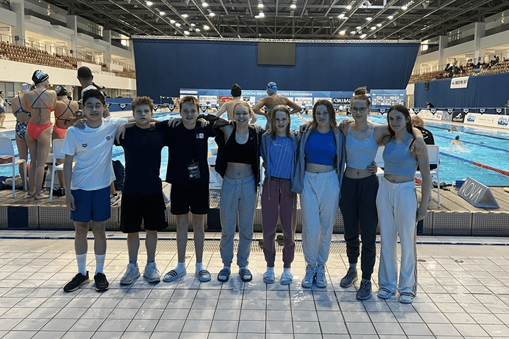 Gruppe von Kindern und Jugendlichen am Beckenrand eines Schwimmwettkampfs in einer Sporthalle.