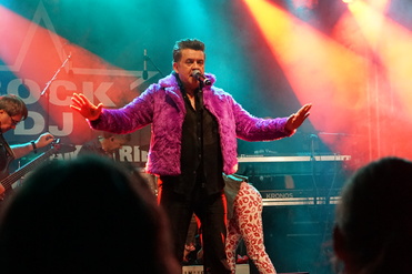 Mann mit lila Pelzmantel singt auf einer Bühne, mit bunten Lichtern und Bandmitgliedern im Hintergrund.