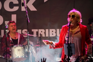 Eine Sängerin in roter Jacke und Sonnenbrille performt auf einer Bühne, während ein Schlagzeuger im Hintergrund spielt.