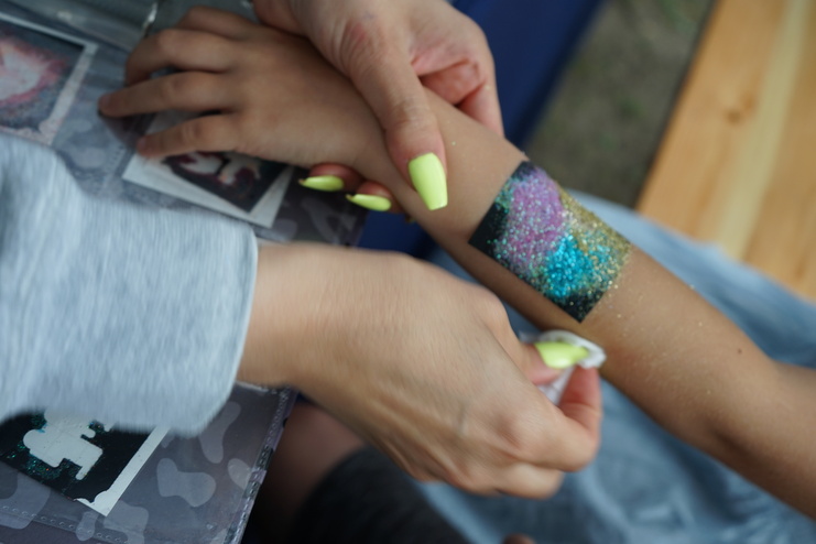 Hände einer Person, die Glitzer auf einen temporären Tattoo-Aufkleber auf dem Arm eines Kindes auftragen.