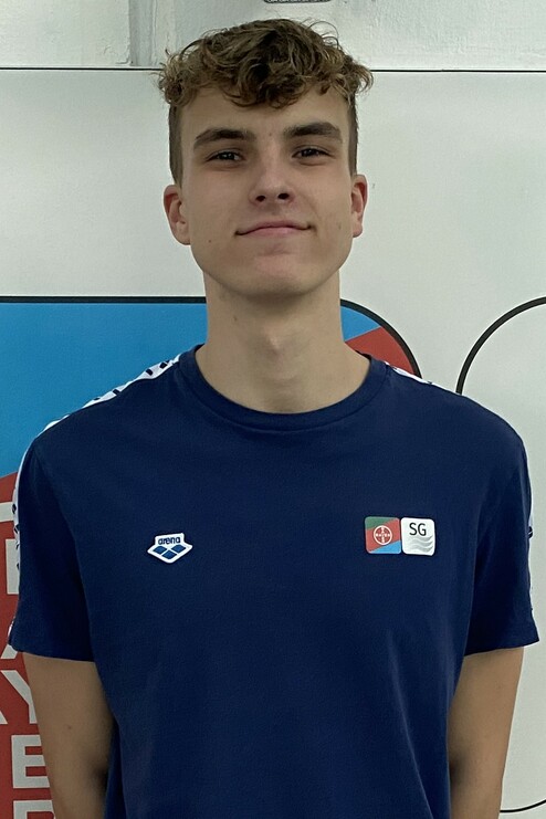 Porträt eines jungen Mannes in einem blauen Shirt mit Logo, vor einer bunten Wand im Hintergrund.