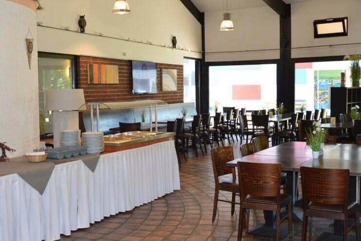 Innenansicht eines Restaurants mit langen Tischen, Stühlen, Buffetbereich und großen Fenstern.