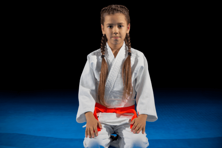 Mädchen in einem weißen Judogi mit rotem Gürtel, kniet auf einer blauen Matte und schaut ernst in die Kamera.