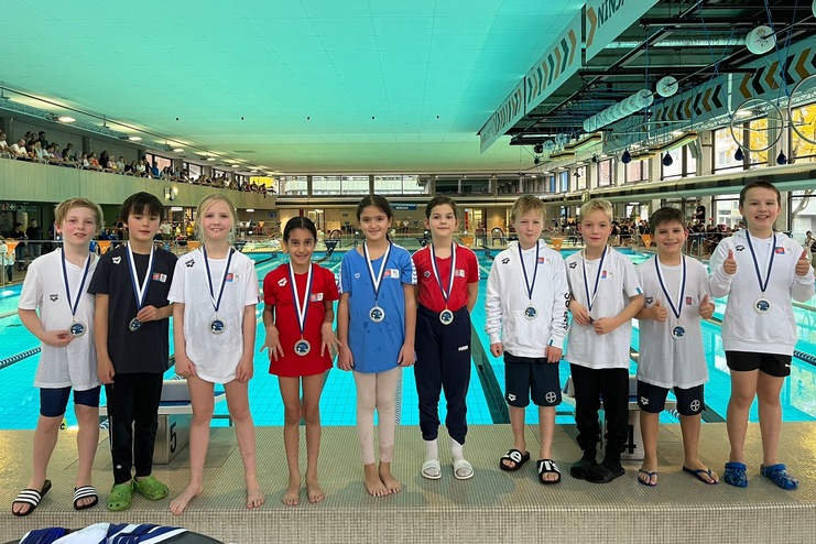 Zehn Kinder in Schwimmbekleidung mit Medaillen stehen vor einem Schwimmbecken in einem Hallenbad.