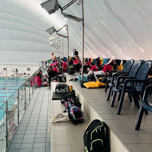 Blick auf eine Tribüne mit Schwimmern, Taschen und Stühlen in einem Schwimmbad unter einem gewölbten Dach.