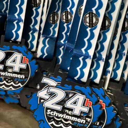 Eine Reihe von Medaillen mit dem Aufdruck „24h Schwimmen“ hängt an blauen Bändern.
