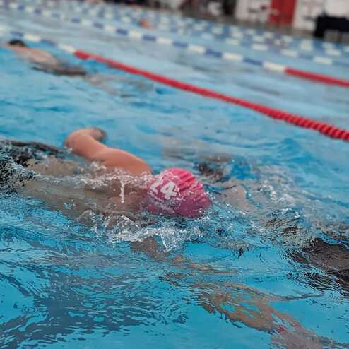 Schwimmerin mit rotem Badekappe schwimmt im blauen Wasser eines Schwimmbeckens. Hintergrund zeigt weitere Schwimmer.