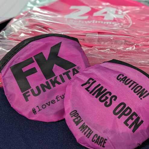 Zwei pinke Schwimmkappen mit dem Aufdruck „FK FUNKITA“ und Warnhinweisen liegen auf einem Tisch, im Hintergrund ein Schwimmbecken.