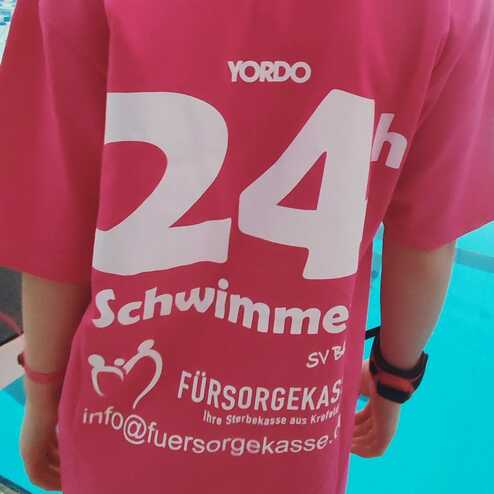 Rückansicht eines Teilnehmers in pinkem Shirt mit der Aufschrift „24h Schwimmen“ am Beckenrand eines Schwimmbads.