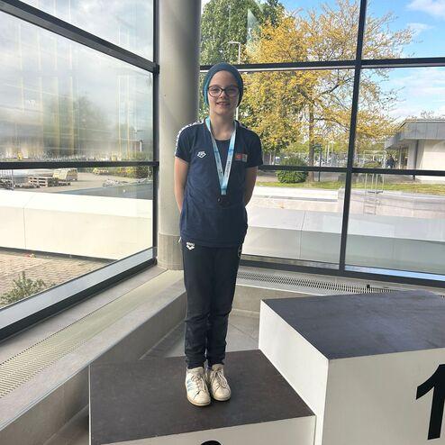 Junge steht auf dem dritten Platz eines Siegertreppchens, trägt eine Medaille und lächelt vor großen Fenstern.