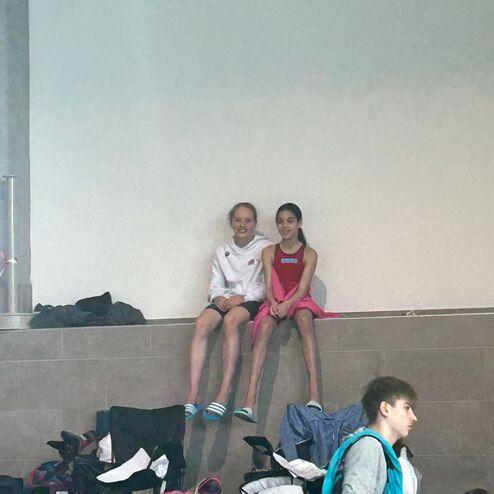 Zwei Kinder in Badebekleidung sitzen auf einer Mauer, umgeben von Schwimmtaschen und Stühlen in einem Schwimmbad.