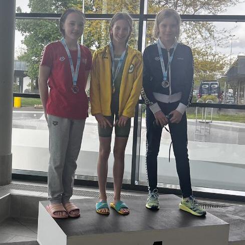 Drei Mädchen auf dem Siegertreppchen mit Medaillen, vor großen Fenstern in einem modernen Gebäude.