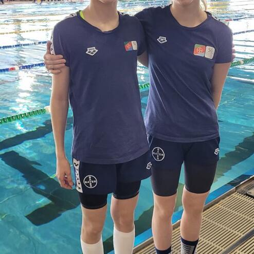 Zwei junge Schwimmerinnen in Vereinskleidung stehen nebeneinander am Schwimmbeckenrand.